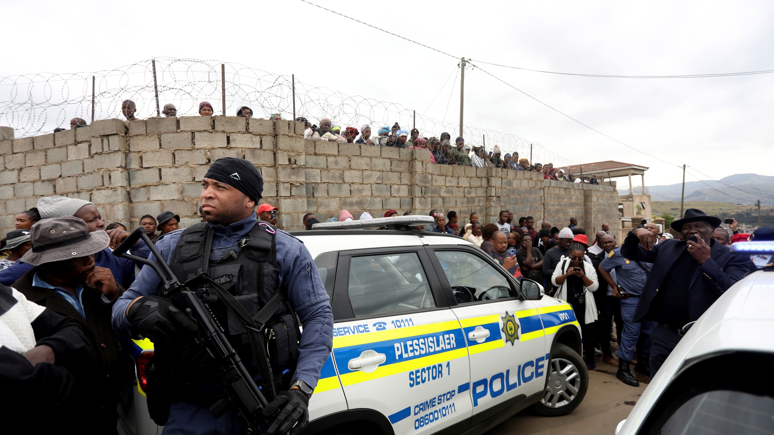 Curious onlookers behind a police cordon, where ten people from the same family were shot dead Friday, April 21, 2023. Police say 10 people were "ambushed" by unknown gunmen at a home in the city of Pietermaritzburg in the eastern KwaZulu-Natal province, South Africa. (AP Photo)