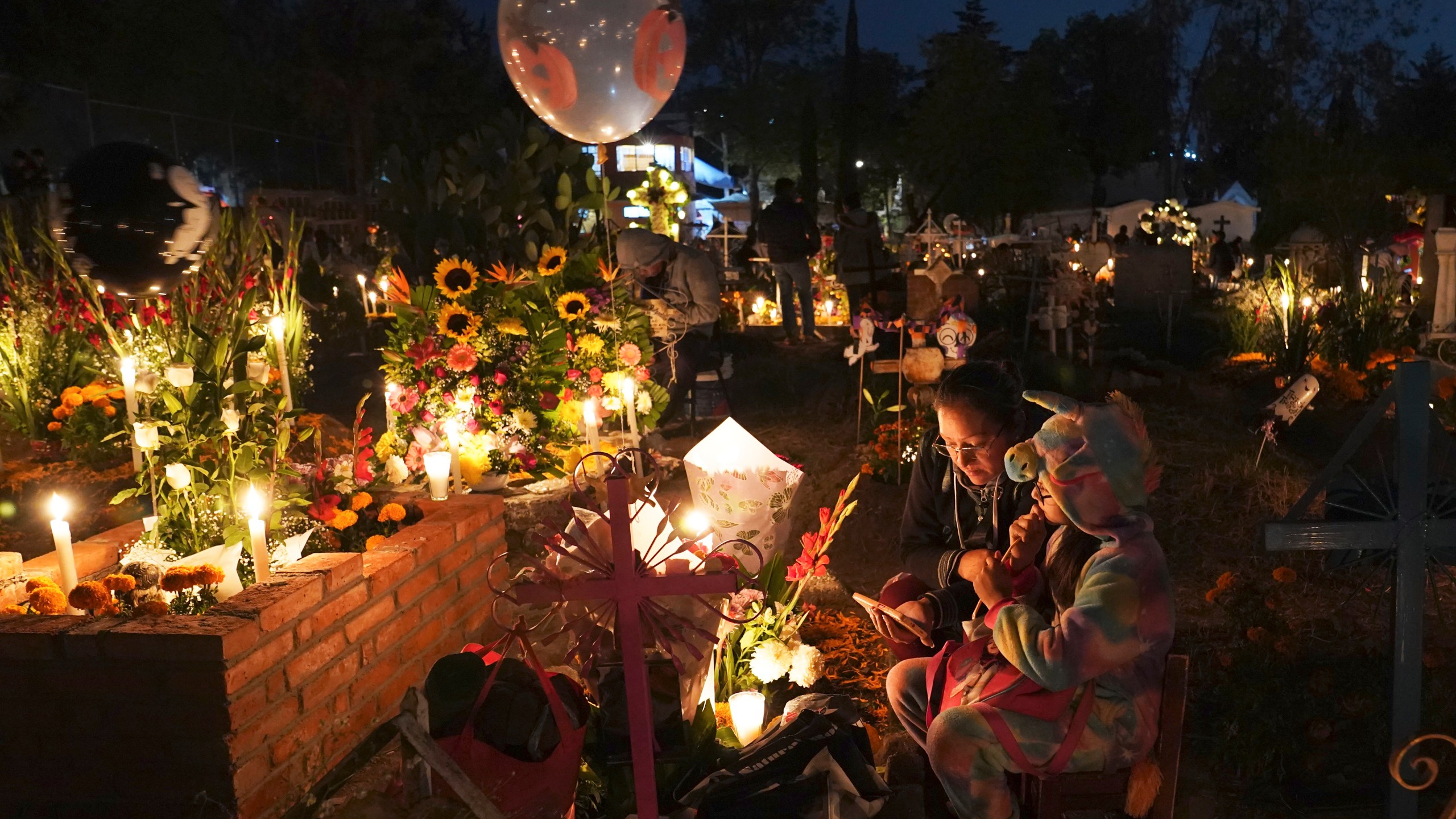 People sit around a child's tomb in the San Gregorio Atlapulco cemetery during Day of the Dead festivities on the outskirts of Mexico City, early Wednesday, Nov. 1, 2023. In a tradition that coincides with All Saints Day on Nov. 1 and All Souls Day on Nov. 2, families decorate graves with flowers and candles and spend the night in the cemetery, eating and drinking as they keep company with their dearly departed. (AP Photo/Marco Ugarte)